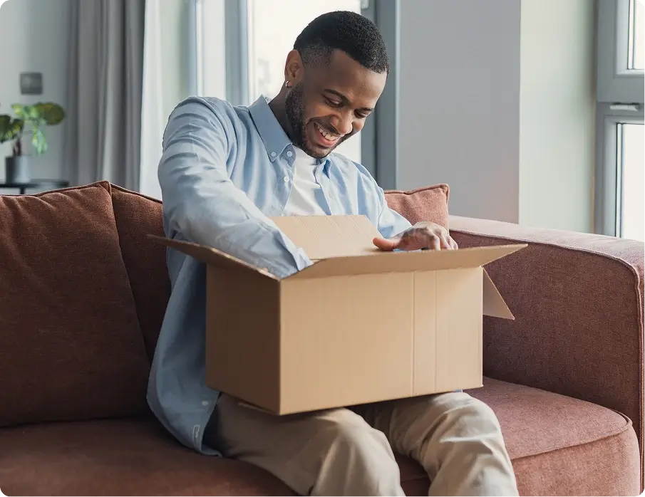 A Black man sitting on a couch opening a box, representing successful order fulfillment with funding support.