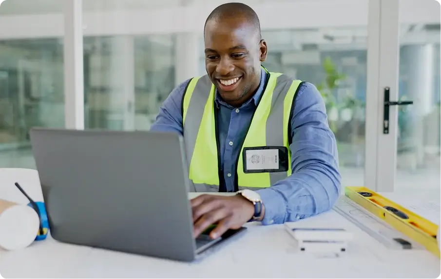 A contract worker in a safety vest smiling and looking at a laptop.