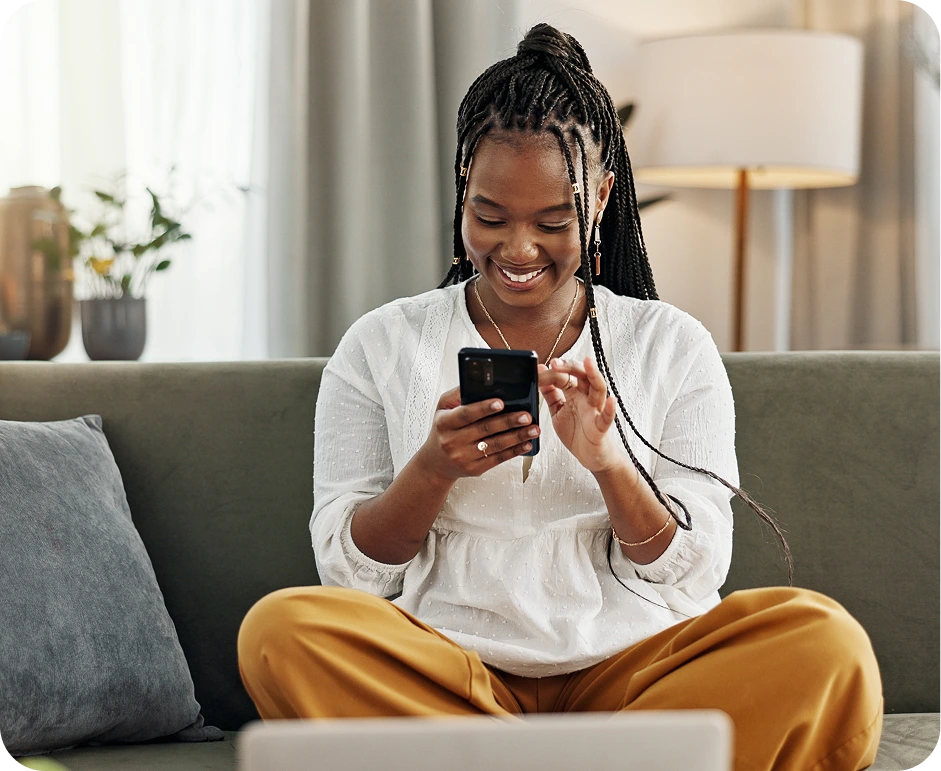A black woman sitting cross-legged on her couch. She is smiling while scrolling on her phone, doing research on Unahina's business funding solutions.
